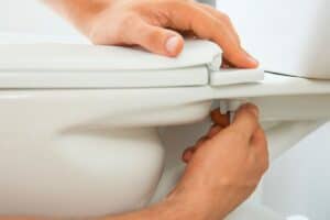 Hands are shown tightening a bolt on the underside of a toilet seat, showcasing a crucial step in the installation or repair process—a handy skill unless you suddenly need an emergency plumber in Madison.