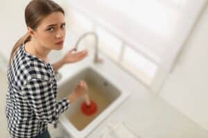 A woman in a plaid shirt is using a plunger on a clogged sink, her eyes reflecting frustration as she considers calling Monroe Emergency Plumbing Repair for help.