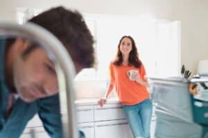 In the spacious, sunlit kitchen, a woman in an orange shirt cradles a mug, her gaze steady and calm. Nearby, a man in a blue shirt stands blurred in the foreground. It's as if they're waiting for the trusted expertise of a 24-hour plumber in Madison to complete their perfect morning.