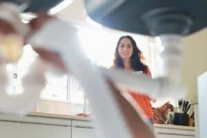 View from under a sink shows a 24-hour plumber in Madison fixing pipes while a smiling woman stands nearby in the kitchen. Bright window light floods the background.