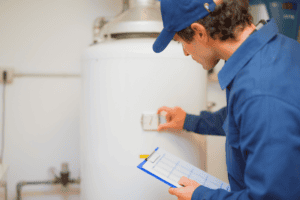 A technician in a blue uniform meticulously inspects a water heater, adjusting settings for optimal performance while holding a clipboard. This expert is well-versed in water heater repairs in Winder, ensuring all adjustments meet the highest standards of efficiency and safety.